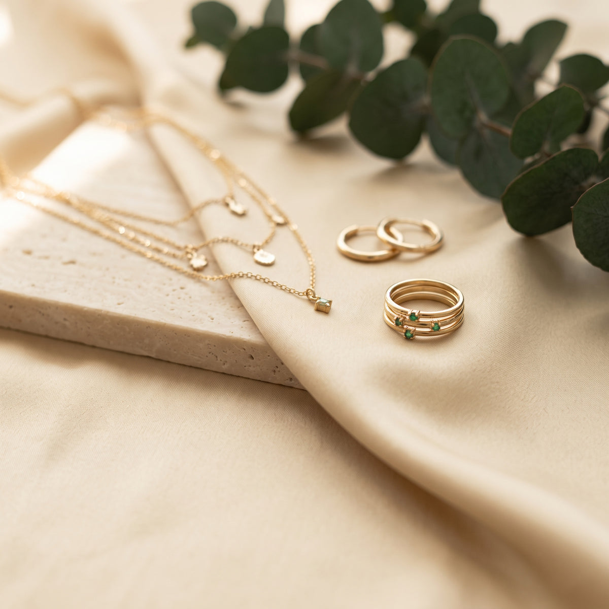Gold necklaces and stacked rings on beige linen and travertine with eucalyptus